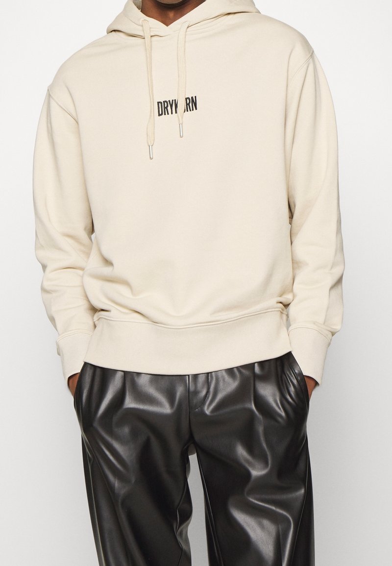 Beżowa bluza z kapturem z kieszenią kangurką z przodu, sznurkiem przy kapturze i czarnym haftem "DRYARN". Połączona z czarnymi, wysokim stanem, spodnie z imitacji skóry.