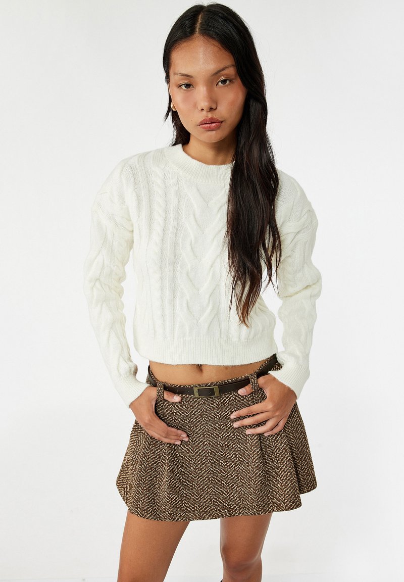 Maglione bianco a maglia intrecciata abbinato a una mini gonna a quadri houndstooth marrone, con una cintura nera. Texture liscia e design attillato.