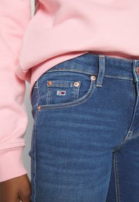 Jeans de mezclilla oscuro con un ajuste ceñido, que presentan un pequeño logo y remaches de cobre. Combinados con un suéter rosa claro. Textura suave.