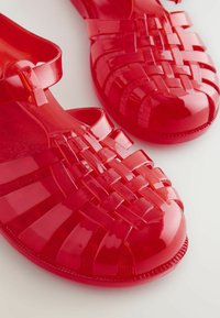 Punane poolläbipaistev plastikust sandaalid, millel on kootud disain, avatud varvas ja pahkluu rihm. Tekstuuriga tallad parandavad haarde ja mugavust.