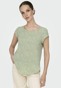 Grünes Kurzarmoberteil mit einem strukturierten, floralem Muster in Off-White. Rundhalsausschnitt mit sanft abgerundetem Saum, kombiniert mit hellen Hosen.