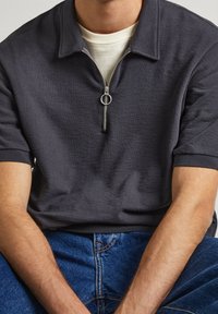 Mörkgrå zip-up polo skjorta med texturerad tyg, korta ärmar och en silverdragkedja. Bärs över en krämfärgad t-shirt.