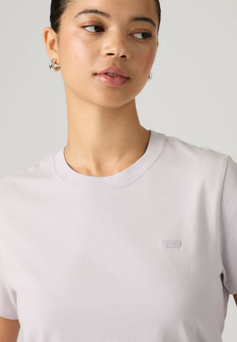 Femme aux cheveux courts portant de petites boucles d’oreilles en forme d’anneau et un t-shirt col rond lavande clair avec un petit logo sur la poitrine.