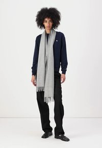 Grijze gebreide sjaal met franjes, om de nek van een persoon die een navy sweatshirt en zwarte broek draagt, in een minimalistische omgeving.