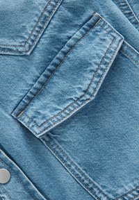 Close-up van een lichtblauwe denim stof met een opvallende zak met zichtbare stiksels en een metalen drukknoopdetail.