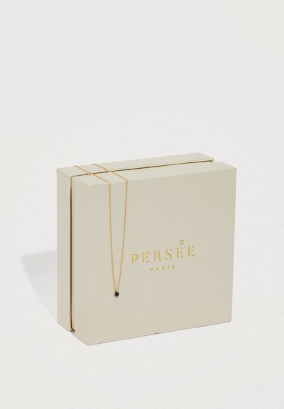 Bēša Perseé Paris rotaslietu kaste ar smalku zelta ķēdīti, uz kuras atrodas neliels melns kulons.