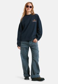 Granatowy sweatshirt z logotypem Roxy, zestawiony z jasnoniebieskimi spodniami o szerokich nogawkach i ciemnozielonymi butami, kontrastujące faktury i kolory.