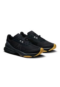 Chaussures de sport noires avec un dessus texturé, des lacets plats et une semelle rembourrée. Elles présentent une semelle extérieure couleur gomme et des accents réfléchissants subtils.