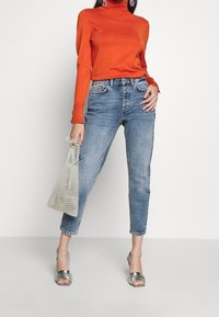 Orange turtleneck sweater, high-waisted light blue jeans, silver high-heeled sandals, clear beaded handbag, casual outfit.
