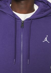 Sudadera con cremallera morada y cordones, logo Jumpman blanco en el lado izquierdo del pecho, llevada sobre una camiseta de cuello redondo blanca.