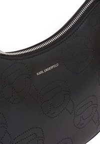 Sac en cuir noir à bandoulière avec un motif texturé de logos graphiques répétés, fermeture éclair argentée et un éclat subtil sur la surface.