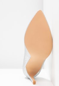 Chaussure à talon haut pailletée argentée avec un bout pointu et une semelle beige lisse, présentant un design élégant et un talon aiguille étroit.