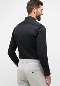 Maglietta nera a maniche lunghe con una texture liscia e polsini con bottone, abbinata a pantaloni grigi chiari con due tasche posteriori e dettagli con bottoni.