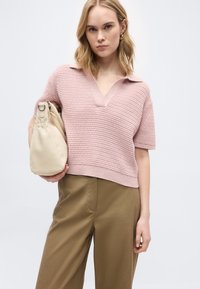 Lyshåret kvinde iført en lyserød strikket kortærmet top og brune bukser, som holder en beige håndtaske mod en ensfarvet hvid baggrund.