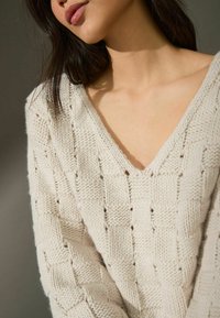 Pull en tricot crème avec un col en V, présentant un motif tissé texturé et des manches amples, mettant en avant un fil épais.