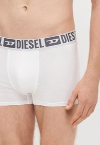 Valged puuvillased bokserpüksid halli elastse vöökohaga, millel on "DIESEL" logo. Suurplaan, mis rõhutab kangast ja sobivust.
