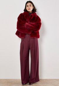 Rote Kunstfelljacke mit lockerer Passform, kombiniert mit bordeauxroten weiten Hosen. Hoher Kragen und reichhaltige Textur. Glänzende Absätze runden den Look ab.