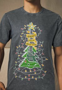 Grå T-shirt med korte ærmer, der har et farverigt grafik af et juletræ og en guitar, med dekorative lyskæder og teksten "Jingle Bell Rock & Roll."