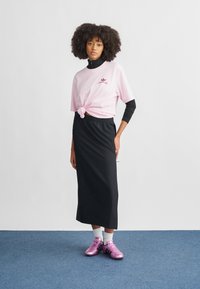 Mulher com cabelo cacheado usando uma camisa rosa com nó sobre uma camisola preta de mangas compridas, saia preta, meias brancas e ténis metálicos cor-de-rosa em tapete azul.