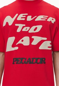 Camiseta de algodón roja con un gran texto gráfico en crema y negro que dice "NUNCA ES TARDE" y "PEGADOR" en diferentes estilos de fuente.