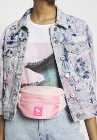 Nerka z jasnoróżowego materiału z dwoma zamykanymi na zamek przegródkami, z metką z logo. Nosi się ją na wielokolorowej koszulce z grafiką i kurtce dżinsowej.