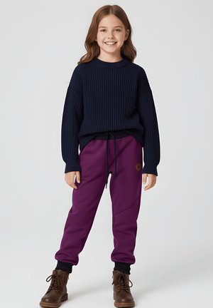 Uśmiechnięta dziewczyna stoi, ubrana w granatowy prążkowany sweter, fioletowe spodnie dresowe z naszywką z logo i brązowe sznurowane buty na jednolitym tle.