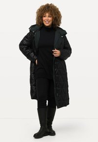 Abrigo largo negro acolchado con capucha y cremallera frontal. Llevado sobre un suéter negro y pantalones, combinado con botas negras hasta la rodilla.