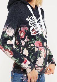 Sudadera azul oscuro con un patrón floral en tonos de rosa y verde, cordones blancos y un bolsillo frontal. Presenta texto de logo en el pecho.