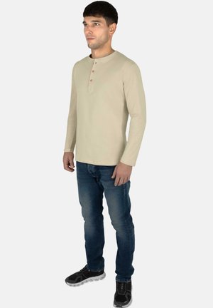 Beige long-sleeve Henley shirt with three buttons at the neck, paired with blue jeans and black athletic shoes. Plain design, minimal detail.