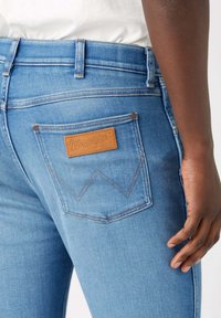 Jeans de mezclilla azul claro con un bolsillo trasero que tiene un parche de cuero marrón y costuras en contraste. El diseño incluye un sutil detalle en forma de Chevron.