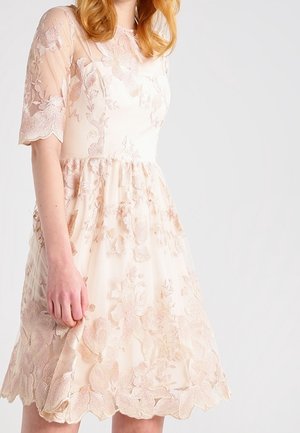 Femme portant une robe rose pâle longueur genoux avec des broderies florales en dentelle et des manches sheer arrivant au coude, sur un fond blanc uni.