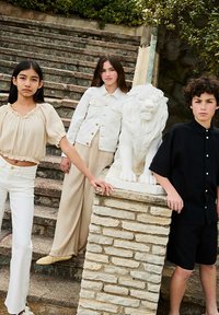 Drie kinderen poseren op stenen trappen naast een witte leeuwstandbeeld. Ze dragen lichte kleding: een beige top, een witte jas en zwarte shorts.