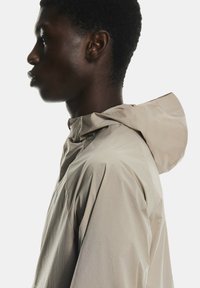 Bēšā vieglā jaka ar kapuci, ar gludu tekstūru un brīvu piegriezumu, nofotogrāfēta no sāniem uz balta fona.