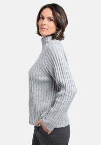 Grauer gerippter Rollkragenpullover mit langen Ärmeln, weichem Material, seitlichen Taschen und tailliertem Design, kombiniert mit dunkelgrauen Hosen.