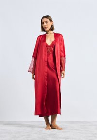 Rød satengkåpe med blondeaksenter på ermene, lagdelt over en matchende satin glidkjole. Glatt tekstur, gulvlengde design.