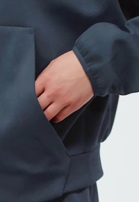 Indumento in tessuto leggero di colore blu scuro con una tasca laterale. Una mano è inserita nella tasca, evidenziando la texture liscia e la vestibilità.