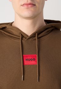 Brun hettegenser med snor. Har et rødt rektangulært merke merket "HUGO." Materialet ser mykt ut med en teksturert overflate.