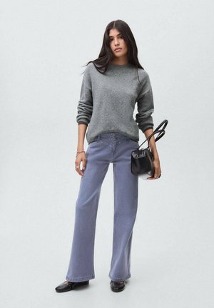 Femme aux longs cheveux noirs, portant un pull gris, un pantalon bleu ample, des chaussures noires et tenant un sac à main noir devant un fond uni.