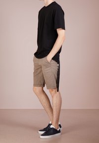 T-shirt en coton noir, shorts beiges avec des manches retroussées et baskets noires. Les shorts comportent des poches latérales et un détail de nœud décoratif.