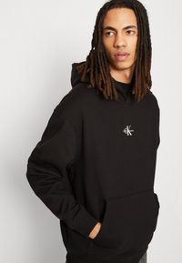Sudadera negra de mezcla de algodón, con un bolsillo delantero, puños acanalados y un pequeño logo en el pecho. Diseño simple y casual.
