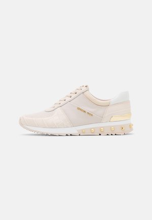 Zapatilla beige de corte bajo con cordones, logo dorado en el lateral, zona de la puntera texturizada y tachuelas cuadradas doradas a lo largo del borde de la suela.