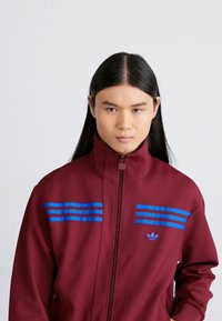 Giacca zip-up bordeaux con colletto alto, caratterizzata da strisce blu sul petto e logo Adidas. Texture liscia, design atletico.