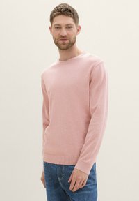 Svetlo roza pulover z dolgimi rokavi iz mehkega in gladkega pletenega materiala z okroglo izrezom in rebrastimi manžetami. Oblečen z modrimi kavbojkami.