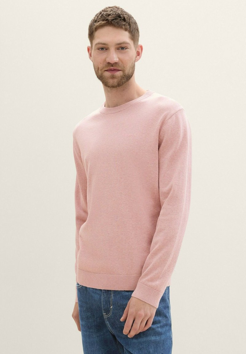 Svetlo roza pulover z dolgimi rokavi iz mehkega in gladkega pletenega materiala z okroglo izrezom in rebrastimi manžetami. Oblečen z modrimi kavbojkami.