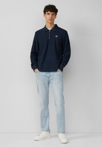 Marineblaues Langarmpolo mit einem Viertel-Reißverschluss und gesticktem Logo, kombiniert mit hellblauen Jeans und weißen Sneakers. Glatter Stoff und entspannte Passform.