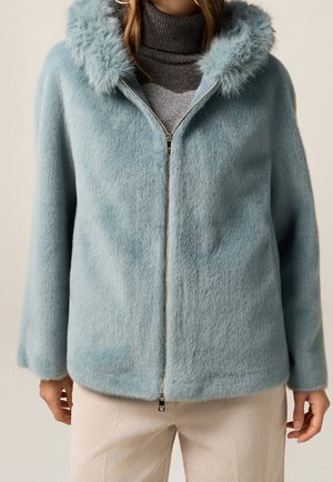 Femme portant une veste légère bleue à fermeture éclair en tissu duveteux, un col roulé gris et un pantalon en velours côtelé de couleur crème.
