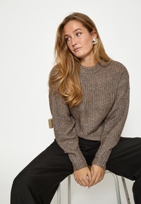 Pruun kudumipullover tekstuuriga mustriga, ringikujulise kaelusega ja pikkade varrukatega. Kombineeritud mustade pükstega. Lihtne disain, minimaalsed aksessuaarid.