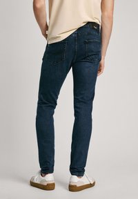 Pantalones vaqueros de mezclilla azul oscuro con un corte ajustado, que presentan dos bolsillos traseros y una etiqueta negra en la cinturilla, combinados con zapatillas blancas.