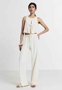 Completo a due pezzi in crema; top corto senza maniche con nodo e dettagli in occhio di pernice, abbinato a pantaloni a gamba larga con dettagli simili.