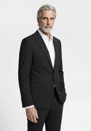 Older man with gray hair and beard wearing a black suit and white shirt standing with one hand in pocket on a plain white background.